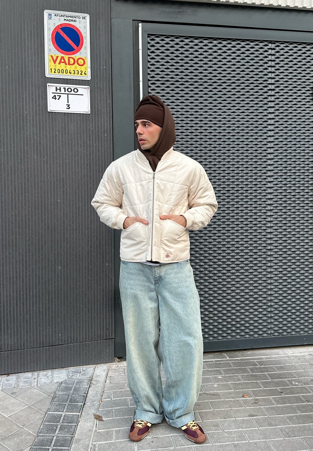 Steppjacke in Cremefarbe, braune Kapuzenjacke darunter, hellblaue, lockere Jeans und braune Sneakers mit gelben Schnürsenkeln, stehend vor einer strukturierten Wand.