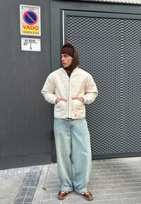 Steppjacke in Cremefarbe, braune Kapuzenjacke darunter, hellblaue, lockere Jeans und braune Sneakers mit gelben Schnürsenkeln, stehend vor einer strukturierten Wand.