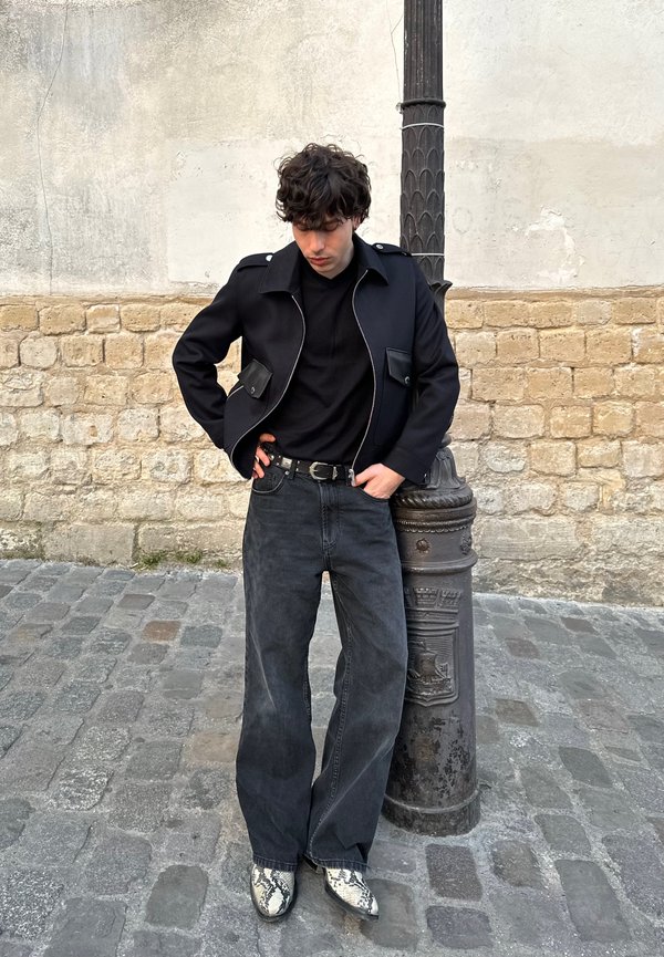 Giubbotto nero con tasche frontali, camicia nera, jeans scuri a gamba larga e stivali con stampa serpente. In piedi accanto a un muro di pietra testurizzato e a un lampione.