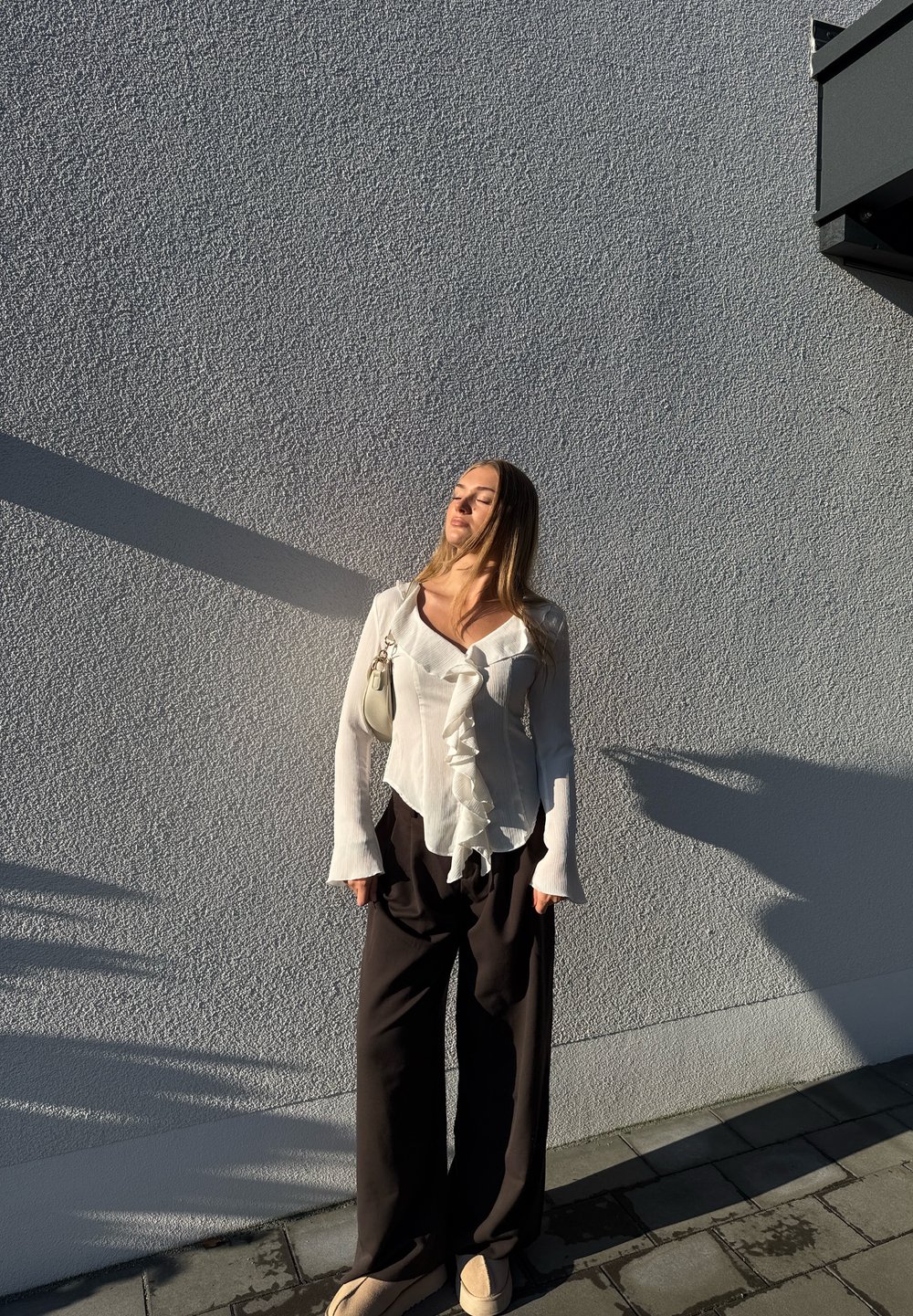 Blusa bianca con ruches e maniche lunghe abbinata a pantaloni neri a gamba larga. Una piccola borsa chiara è appesa a una spalla.