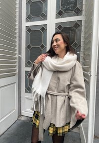 Μπεζ μαλλίνα παλτό με φαρδιά μανίκια, ζώνη στη μέση και ένα χοντρό άσπρο κασκόλ με φούντες. Το φόντο έχει σχέδια από γυάλινες πόρτες και παντζούρια.