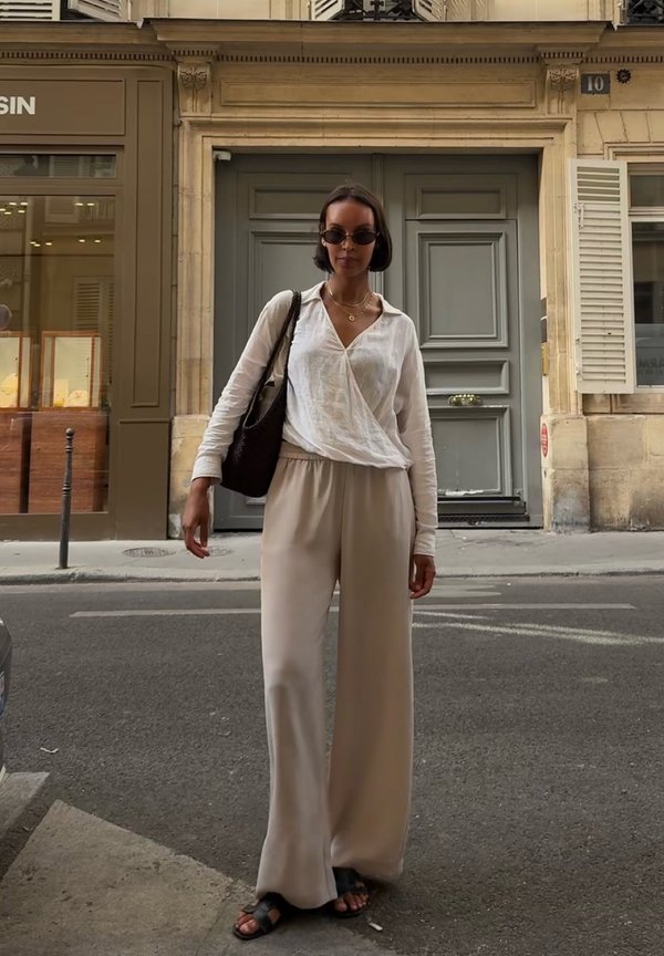 Mujer con gafas de sol, blusa blanca, pantalones beige de pierna ancha y sandalias negras, de pie en una calle de la ciudad cerca de una puerta gris.