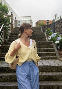 Cardigan canela clara com textura e fecho de botão sobre uma t-shirt branca, combinado com calças riscas azuis claras e uma bolsa de palha, em escadas de pedra.