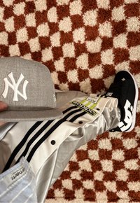 Szara czapka New York Yankees, błyszczące srebrne spodnie z czarnymi paskami i czarne trampki DC z białym logo, na strukturalnym pomarańczowo-beżowym dywaniku.
