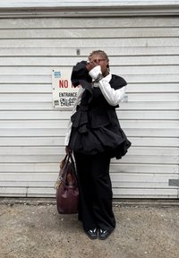 Rochie neagră în straturi cu volane peste o cămașă albă, pantaloni negri și o geantă burgundy. Include accesorii metalice și pantofi cu model.