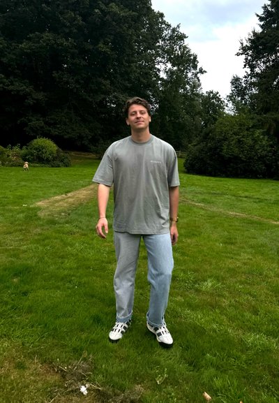 Camiseta de gran tamaño gris, vaqueros azul claro y zapatillas blancas con detalles en negro, de pie sobre hierba verde en un entorno de parque.