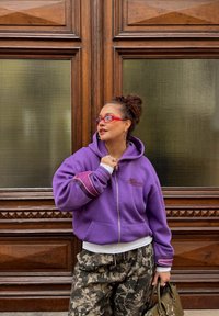 Sweat à capuche violet à fermeture éclair avec un logo, chemise blanche superposée, pantalon baggy camouflage et sac vert. Les accessoires comprennent des lunettes rouges et des boucles d'oreilles.