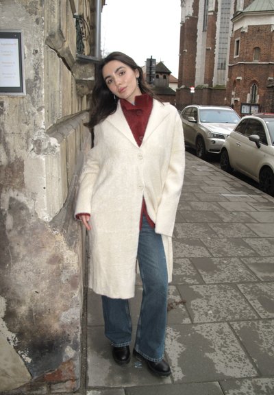 Joven mujer de cabello oscuro se apoya contra una pared de piedra desgastada en una calle de la ciudad, vistiendo un abrigo crema, suéter rojo, jeans azules y zapatos negros.