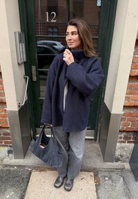 Velik plašč iz mornarsko modre volnene mešanice z visokim ovratnikom in gumbi, kombiniran z sivimi hlačami z navpičnimi črtami in črnimi škornji.