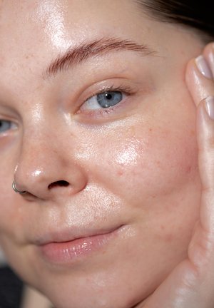 Primo piano di un volto con pelle chiara, caratterizzato da una texture lucida e idratata. Leggeri efelidi, occhi azzurri e un anello al naso visibili.