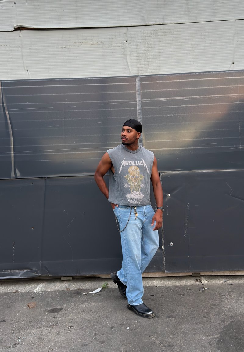 Homme portant un débardeur Metallica, un jean bleu clair, des chaussures noires et une coiffe noire, debout contre un mur métallique gris en extérieur.