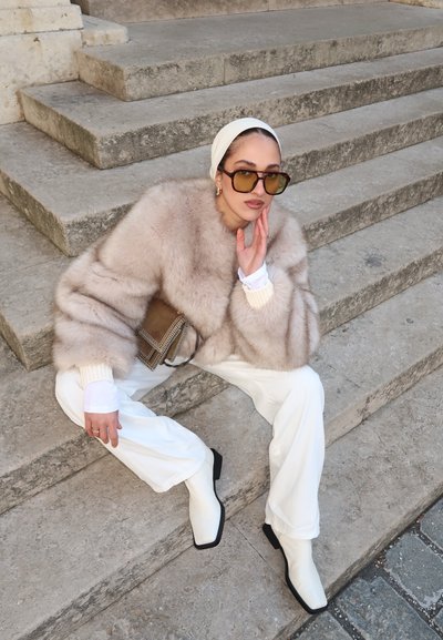 Chaqueta de piel sintética en beige claro, pantalones de pierna ancha blancos y botas tobilleras blancas con ribetes negros. Gafas de sol marrones de gran tamaño y un bolso beige.