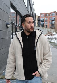 Homme aux cheveux courts et foncés et à la barbe portant une veste beige sur un pull noir, debout à l'extérieur près de bâtiments modernes par une journée nuageuse.