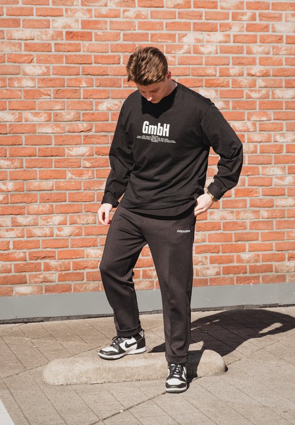 Camisa negra de manga larga con el logo y texto "GmbH", combinada con pantalones de chándal negros con el texto "PECADOR". Se lleva con zapatillas negras y blancas.