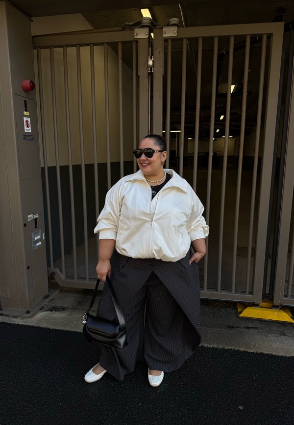 Woman wearing sunglasses, a white jacket, wide pinstriped pants, and white shoes stands smiling, holding a black handbag near a metal gate.