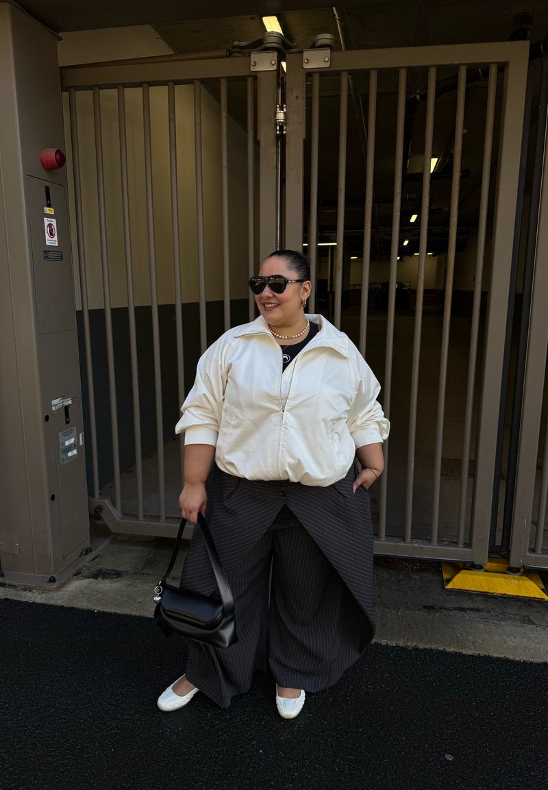 Femme portant des lunettes de soleil, une veste blanche, un pantalon large à rayures et des chaussures blanches, debout en souriant, tenant un sac à main noir près d'une porte en métal.