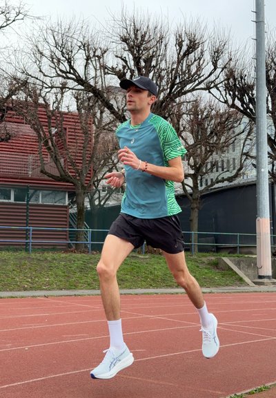 Hombre con gorra, camiseta azul verdosa y verde, pantalones cortos negros y zapatillas blancas corriendo en una pista al aire libre con árboles sin hojas al fondo.