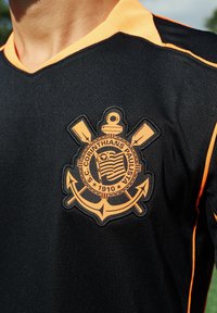 Černý sportovní dres s oranžovými detaily, na kterém se nachází texturované vyšívané logo kotvy a zkřížených pádel, označený "S.C. Corinthians Paulista 1910."