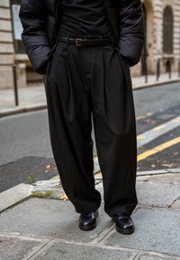 Pantalon noir plissé en tissu léger, porté avec une ceinture noire et des chaussures noires brillantes, sur fond urbain.