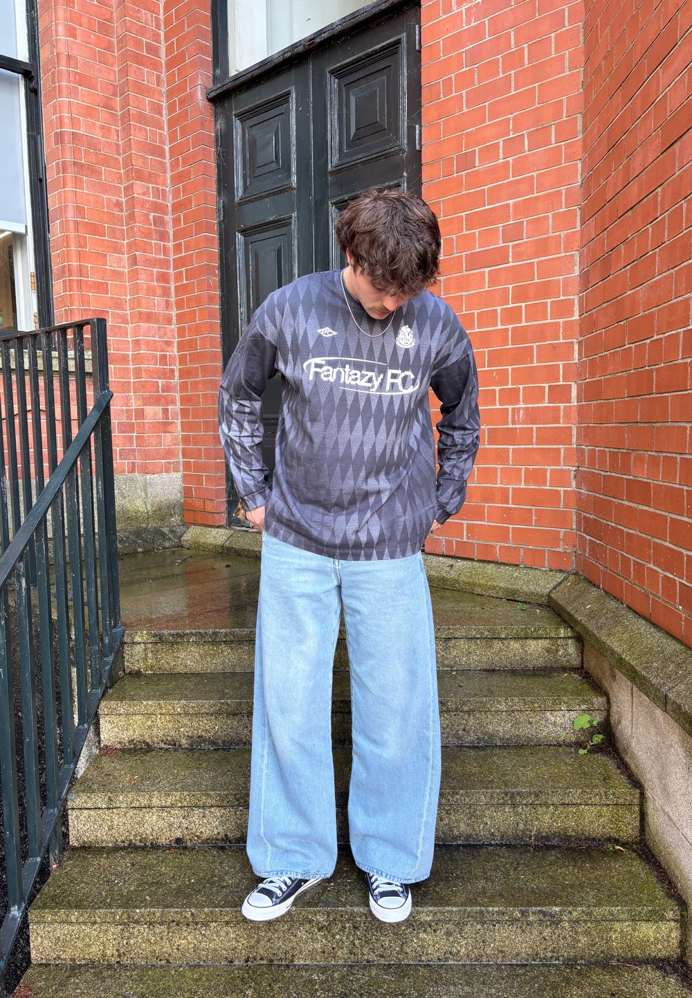 Long-sleeved black and grey diamond-patterned shirt featuring the "Fantasy FC" logo, teamed with wide light blue jeans and black-and-white trainers.