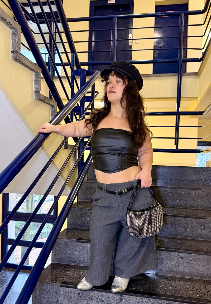 Schwarzes Leder-Crop-Top, graue weite Hose mit Gürtel, silberne Schuhe, strukturierte schwarze Tasche, stehend auf einer Treppe mit blauen Geländern.