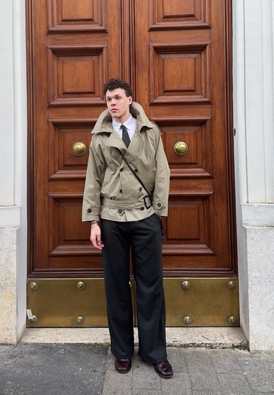 Joven con gabardina beige, camisa blanca, corbata negra, pantalones anchos negros y zapatos oscuros, de pie frente a una gran puerta de madera con detalles de latón.