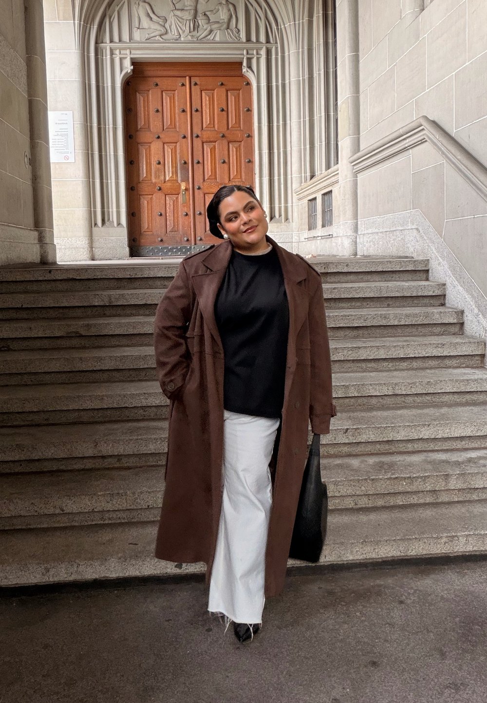 Ilgas rudas paltas ant juodo viršutinio drabužio, baltos platėjančios kelnės ir juoda rankinė. Stovi ant akmeninių laiptų, fone matyti medinės durys.