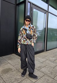 Casaco de fleece colorido com padrão e gola azul, mochila preta, calças largas pretas e sapatos pretos. Óculos de sol e um gorro completam o visual.