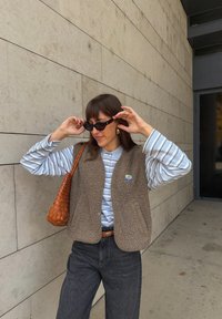 Brūns flīsa veste virs gaiši zila svītrainā krekla, kombinēta ar melnām platām biksēm un austu brūnu somu. Valkā melnas saulesbrilles.