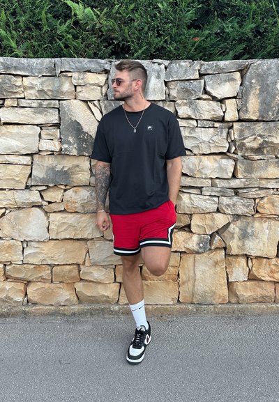 Camiseta negra de algodón, pantalones cortos rojos con rayas negras y blancas, zapatillas negras, de pie contra una pared de piedra, usando gafas de sol.