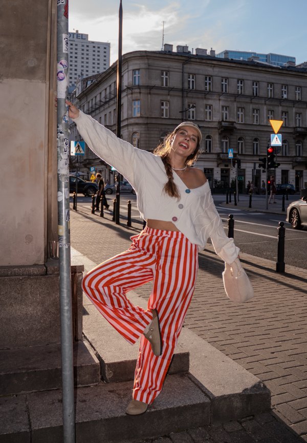 Suéter cropped branco com botões, calças listradas vermelho e branco, e sapatos bege. Ambiente urbano bem iluminado com edifícios ao fundo.