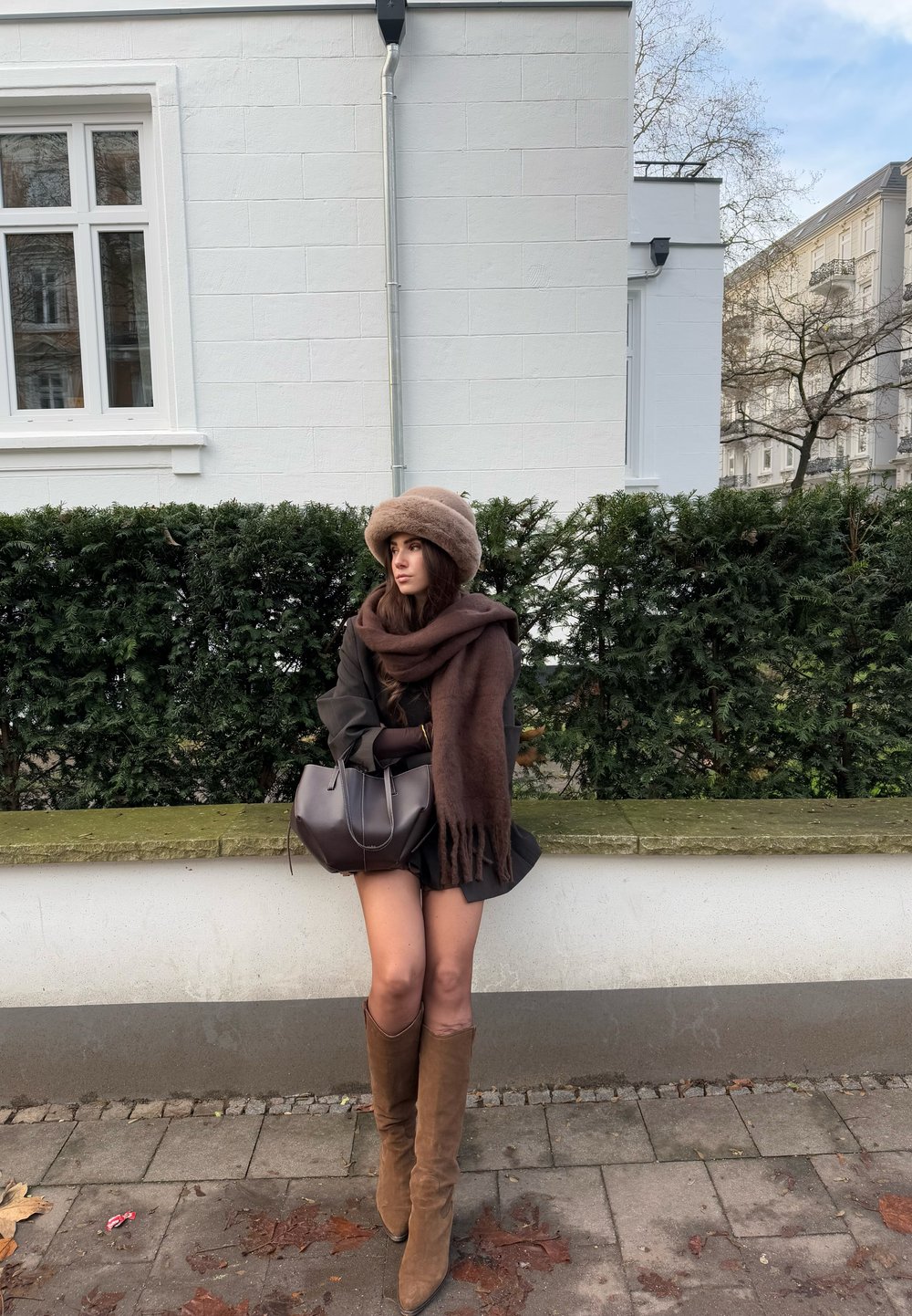 Brūns šalle un cepure, tumša apģērba virsdaļa un līdz ceļiem augsti beidži zābaki. Tur rokās tumšu rokassomu. Persona sēž uz akmens sienas ar zaļumu aiz muguras.