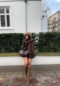 Brūns šalle un cepure, tumša apģērba virsdaļa un līdz ceļiem augsti beidži zābaki. Tur rokās tumšu rokassomu. Persona sēž uz akmens sienas ar zaļumu aiz muguras.