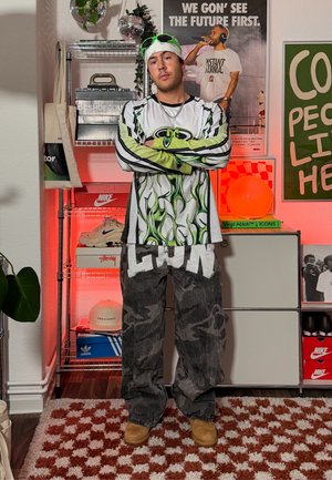 Hombre con camisa gráfica de manga larga y pantalones anchos está de pie con los brazos cruzados en una habitación con estantes, zapatillas, plantas y pósters visibles detrás de él.