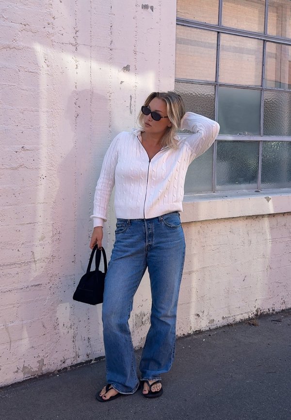 Mujer con gafas de sol, llevando un suéter blanco, vaqueros azules y sandalias negras, sosteniendo un pequeño bolso negro y de pie contra una pared blanca de ladrillos.