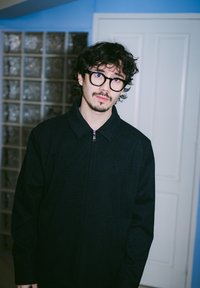 Črna jakna z zadrgo s teksturiranim vzorcem, visokim ovratnikom in dolgimi rokavi. V ozadju prozorna steklena opečna stena in bela vrata.