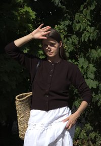 Mørkebrun cardigan med knapper, paret med et hvitt skjørt med blonder. En vevd stråveske hviler på den ene skulderen, mot en bakgrunn av grønt.