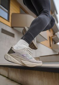 Chaussure de course gris clair avec dessus texturé, accents réfléchissants et semelle épaisse à motifs. Portée avec des chaussettes blanches aux chevilles et des leggings noirs.