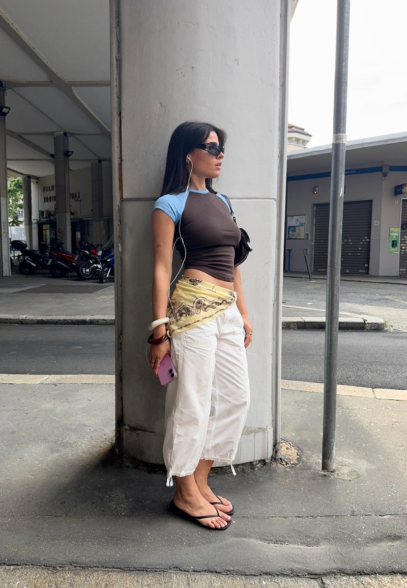Femme portant des lunettes de soleil, des écouteurs, tenant un téléphone, appuyée contre une grande colonne blanche sur un trottoir en ville.