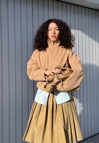 Veste beige zippée, chemise bleu clair en dessous, jupe kaki plissée, et sac en cuir à la main. Cheveux bouclés et fond neutre.