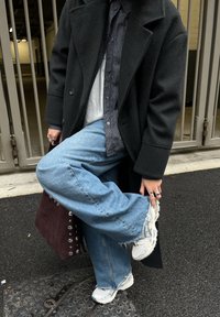 Personne portant un manteau foncé, une chemise à carreaux, un jean large bleu, des baskets blanches, tenant un pied et portant un sac à main bordeaux clouté.