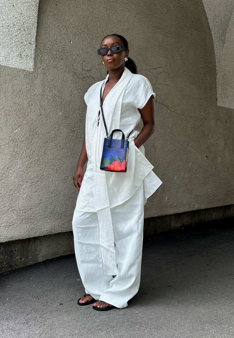 Outfit in lino bianco con maniche corte, vita cinte e pantaloni a gamba larga. Piccola borsa nera con un design floreale colorato. Occhiali da sole neri.