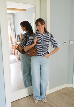 Mujer con cabello castaño, con una blusa blanca y negra de cuadros y vaqueros azul claro, está de pie sonriendo y sosteniendo un bolso marrón junto a un espejo de cuerpo entero en el interior.