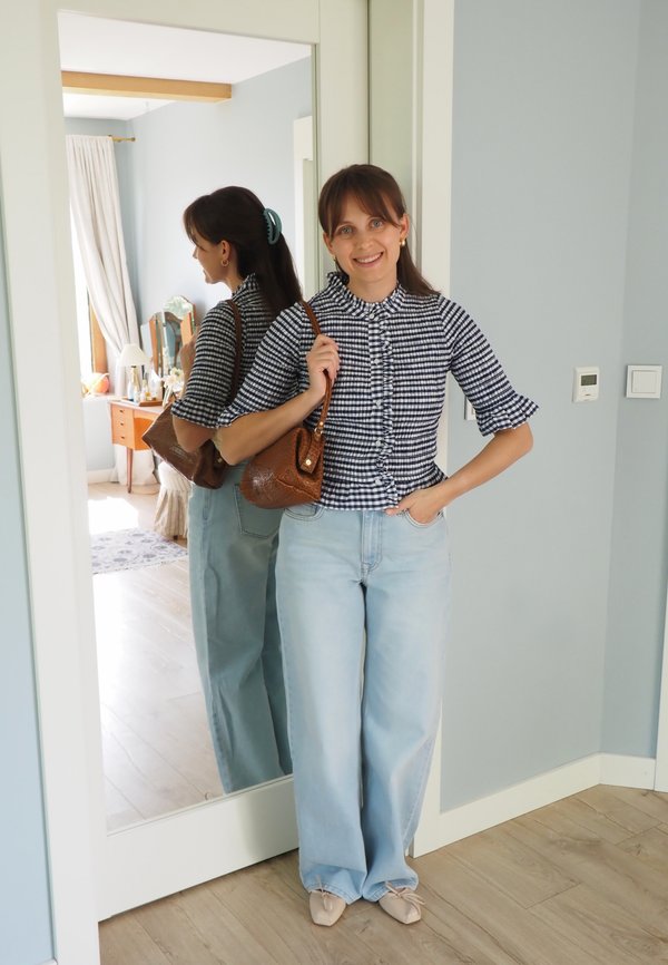 Žena s hnědými vlasy v černo-bílé kostkované blůze a světle modrých džínách stojí usměvavá, drží hnědou kabelku u zrcadla na celou délku v interiéru.