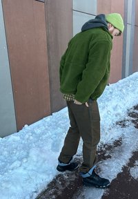 Homme portant une veste verte et un bonnet, un pantalon marron, des bottes noires, debout sur un sol enneigé à côté d'un mur à panneaux multicolores.