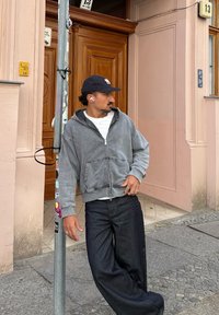 Grey zip-up hoodie with a large hood, paired with loose dark denim jeans. The model wears a navy cap and white earbuds, leaning against a pole.