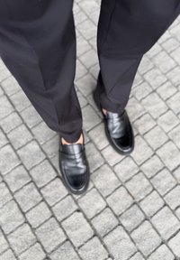 Zwarte leren loafers van glad leer, met een ronde neus en een lage hak, gecombineerd met een op maat gemaakte zwarte broek, op een kinderkopjesoppervlak.