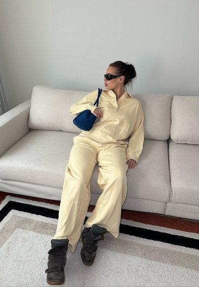 Mujer con conjunto beige y gafas de sol negras sentada en un sofá gris claro sosteniendo un pequeño bolso azul, mirando hacia su izquierda.