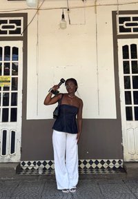 Top senza spalline in denim, pantaloni bianchi a gamba larga, occhiali trasparenti. Regge un telefono vintage, in piedi contro un muro semplice con piastrelle da pavimento a fantasia.