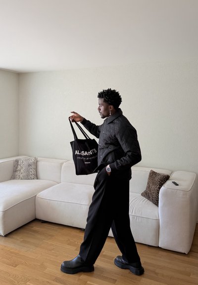 Hombre con chaqueta acolchada negra y pantalones caminando en interiores sosteniendo una bolsa tote negra de AllSaints junto a un sofá seccional blanco con cojines.